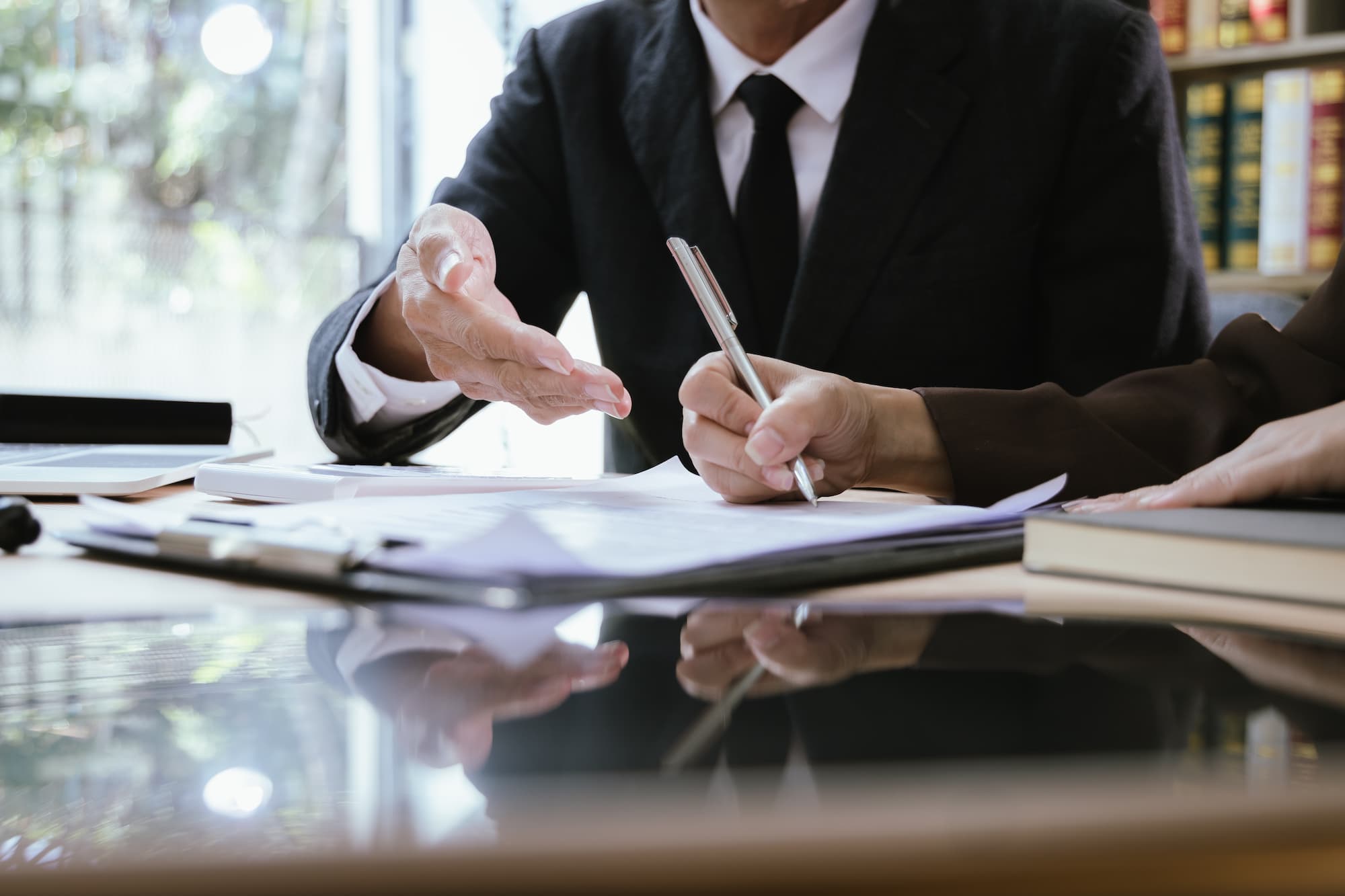 Close-up of an attorney in a black suit and tie gesturing with an open hand toward a document on a clipboard while a client in a brown jacket signs it with a silver pen. The papers rest on a reflective desk beside a laptop and a hardcover book, with a bright window on the left and shelves of law books in the background.