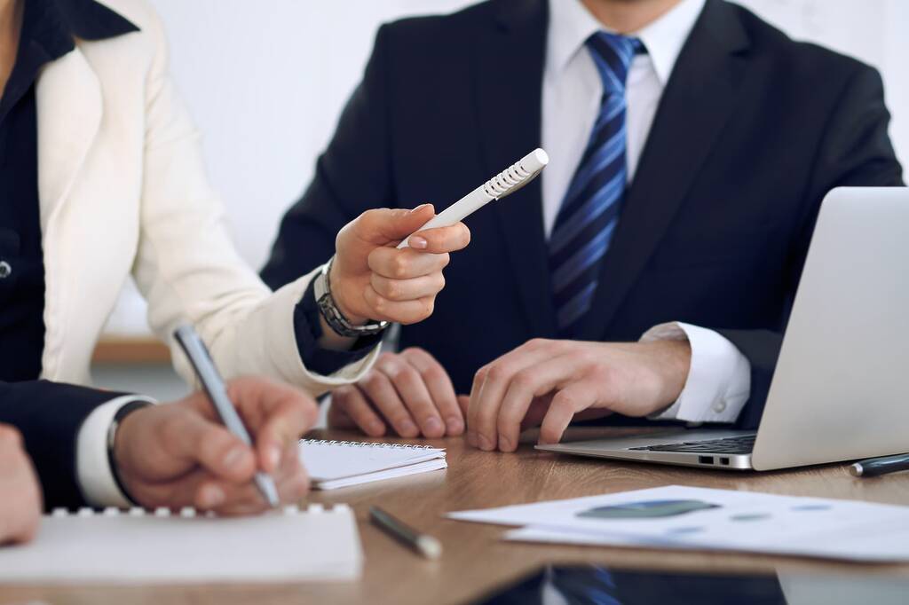 Close-up of three professionals at a wooden conference table. A woman in a cream blazer gestures with a white pen toward a man in a dark suit and blue striped tie, whose hands rest beside an open laptop, while a third person in the foreground writes in a spiral notebook. Printed documents and pens are spread across the table.