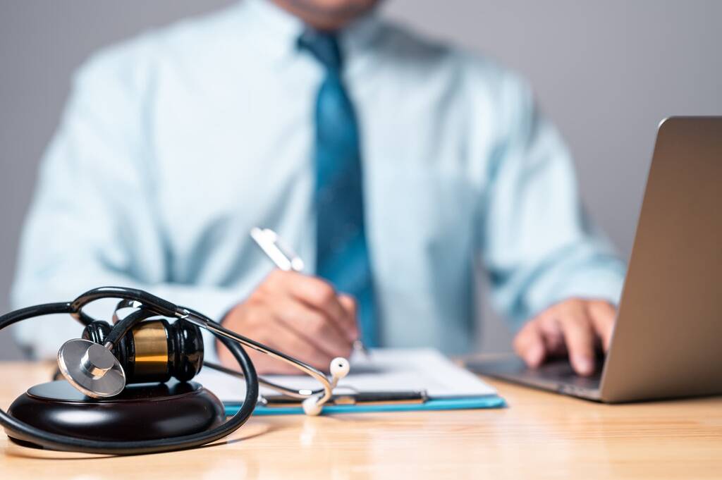 Close-up of a stethoscope wrapped around a wooden judge's gavel and sound block on a light wood desk. In the soft-focus background, a professional in a light blue shirt and teal tie writes on a clipboard with one hand while typing on a laptop with the other, suggesting legal work on a personal injury or medical case.