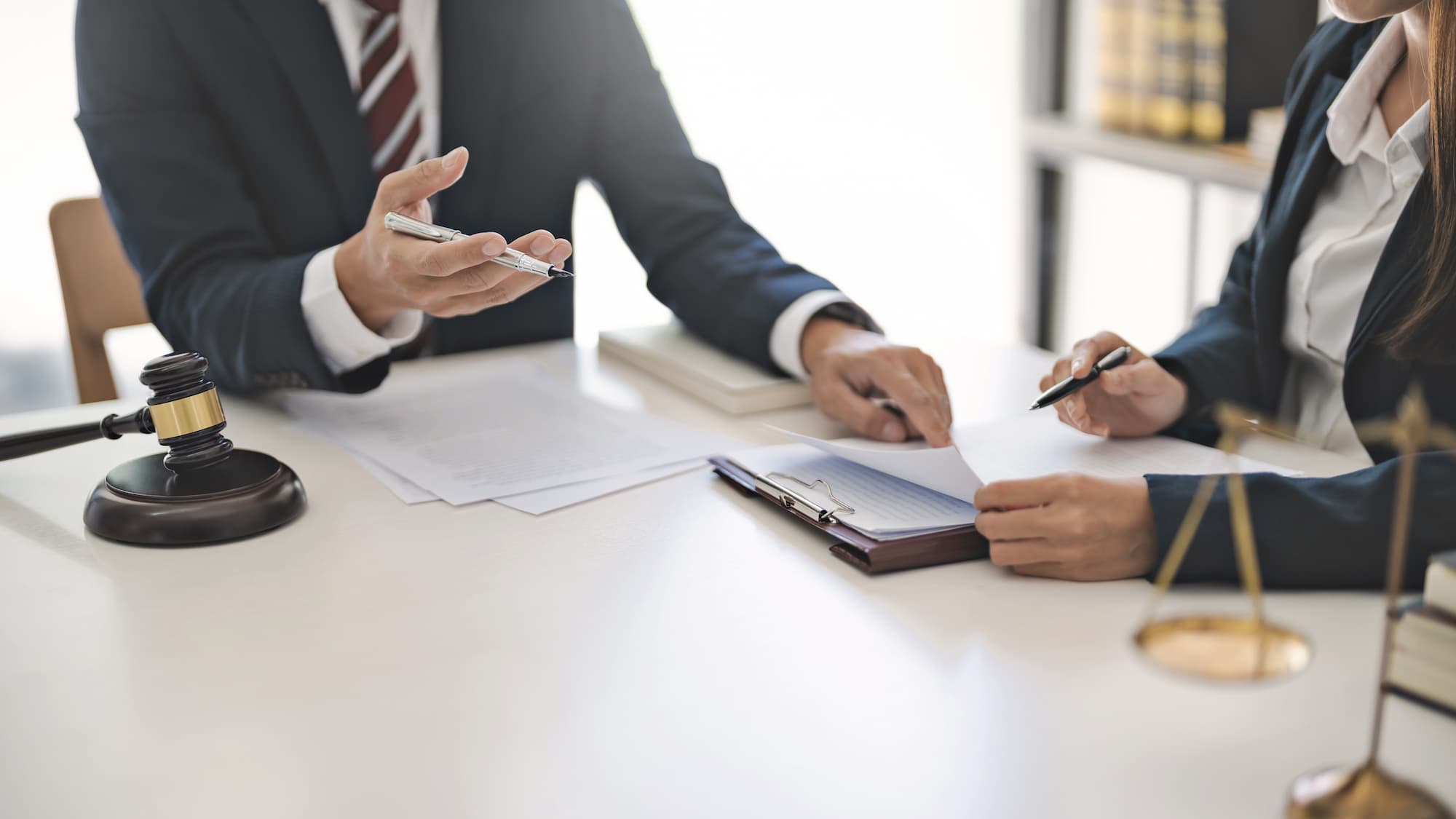 Two professionals in dark suits sit at a bright white desk reviewing legal documents together. One gestures with a silver pen while pointing to a page on a clipboard, as the other holds a pen and turns the paper. A wooden judge's gavel rests on the left, brass scales of justice sit on the right, and law books line a shelf in the soft-focus background.